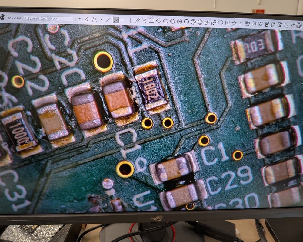Close-up of a circuit board on a monitor screen, showing resistors, capacitors, and copper traces.
